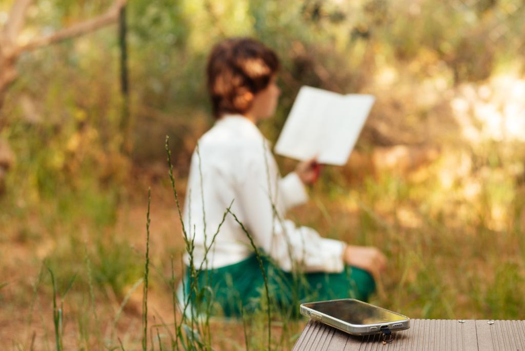 Eine Frau liest ein Buch draußen in der Natur. Im Vordergrund ein Handy auf einem Tisch.