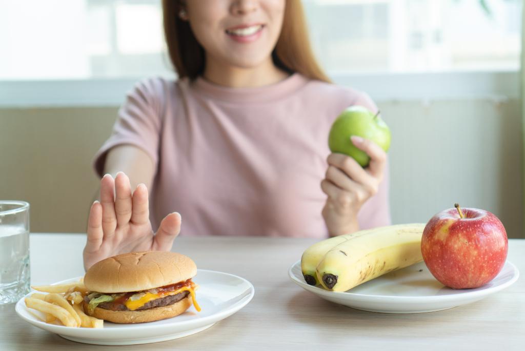 Eine Frau mit Apfel in der Hand schiebt einen Teller Junk-Food von sich weg.