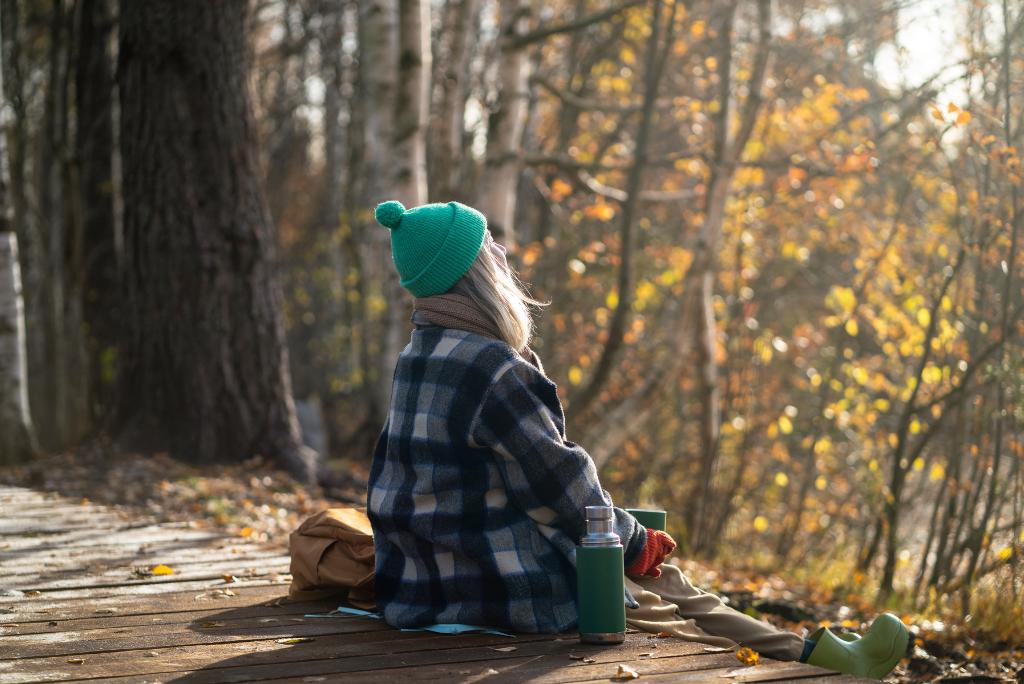 Eine Frau im Wintermantel sitzt mit Thermoskanne draußen in der Sonne.