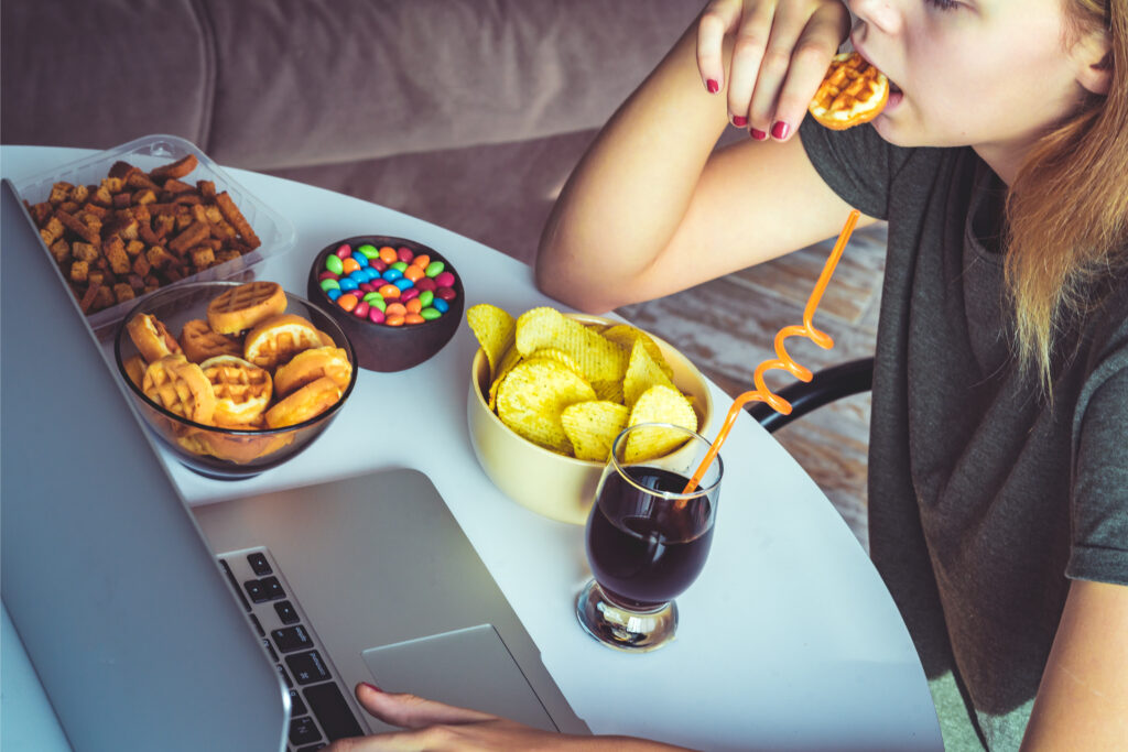 Eine Frau sitzt vor ihrem Laptop und isst dabei ungesunde Snacks.